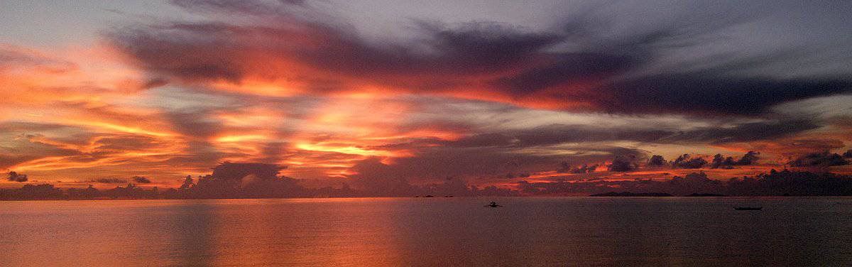 Vibrant Palawan sunset over a calm sea, with orange and purple clouds reflected on the water and small boats from island hopping adventures dotting the horizon.