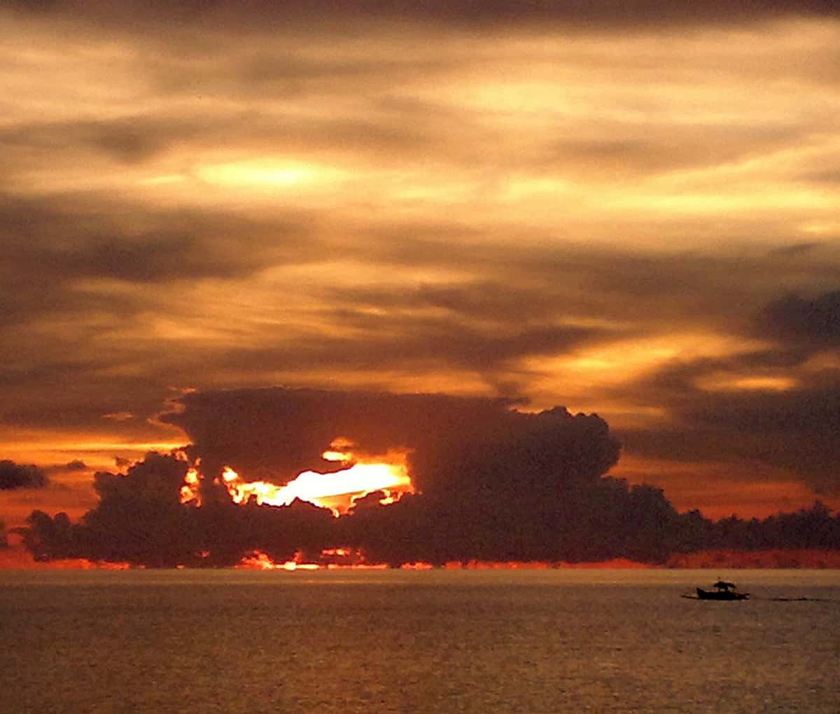 A dramatic sunset over the ocean, with dark clouds casting shadows and an orange glow painting the sky, highlights a small boat silhouetted on the water—a perfect scene for a serene boat tour.