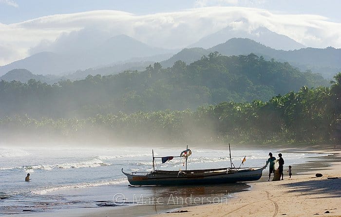 A boat, ready for an island hopping adventure, sits on a misty beach with majestic mountains in the background and curious onlookers standing nearby.