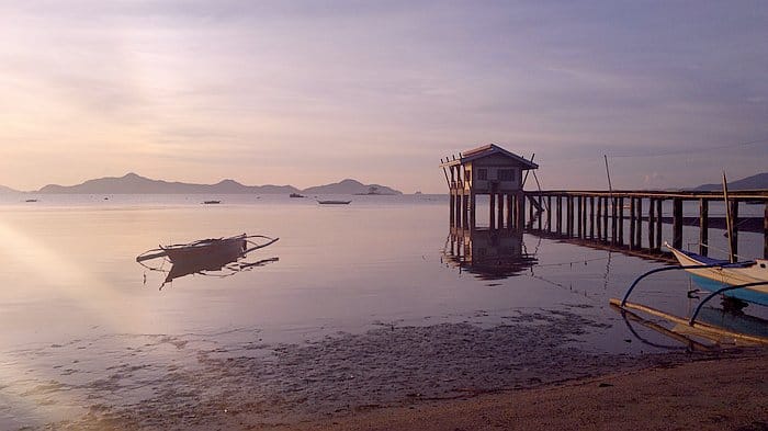 Tranquil seaside scene with a wooden pier, a small house on stilts, distant mountains, and a traditional boat perfect for island hopping on calm water at sunset.