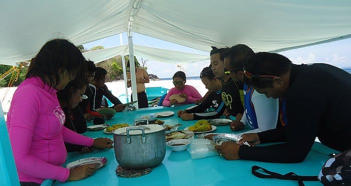 People in wetsuits sit around a turquoise table on a boat tour, enjoying a meal with various dishes while island hopping.