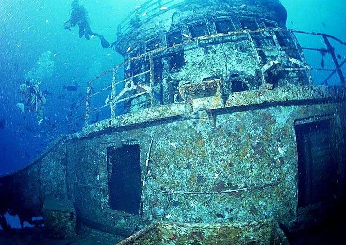 Divers, fresh off an exciting island hopping adventure, explore a large, coral-encrusted shipwreck resting majestically on the ocean floor.