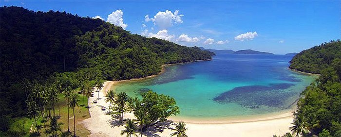 Aerial view of a tropical bay with lush green hills, turquoise water, and a sandy beach lined with palm trees, perfect for island hopping under a partly cloudy sky.