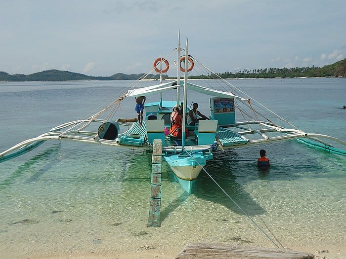 A blue and white outrigger boat docked on a sandy beach with clear, shallow water is perfect for island hopping in the Philippines. Three people are aboard, while one stands in the water near the boat. Hills and greenery provide a scenic backdrop under a cloudy sky.