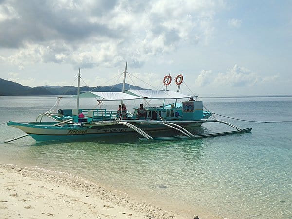 A turquoise outrigger boat is anchored on a clear, shallow shoreline. Two people sit under a canopy, poised for island hopping. The backdrop features the tranquil sea stretching towards the distant mountains of the Philippines under a partly cloudy sky.
