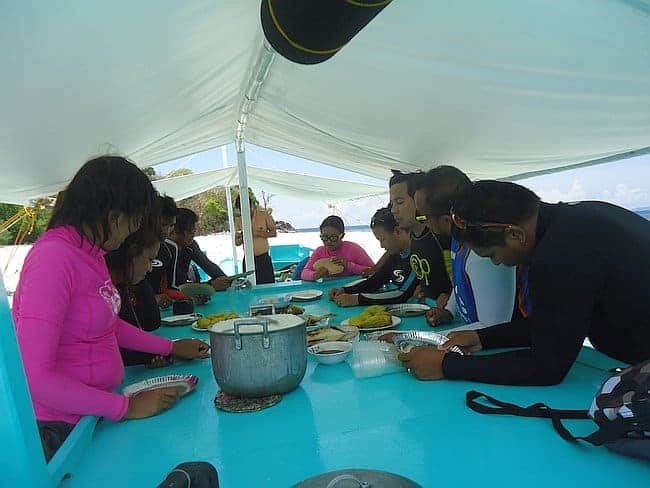 A group of people in swimwear are seated around a table on a boat under a canopy, sharing a meal after an exciting day of island hopping. The table is brimming with dishes served in various sizes. It's a sunny day with clear skies and the ocean glistening against the backdrop of the Philippines.