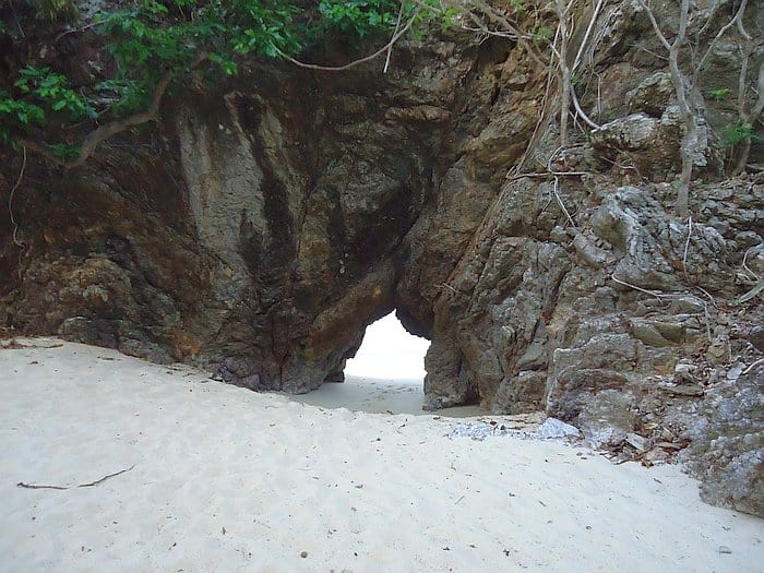 A natural rock archway on a sandy beach in Palawan offers an enchanting view. The rocky formation, surrounded by lush greenery, serves as a perfect backdrop for island hopping adventures, with the archway leading to the ocean beyond, framed by rugged rocks.