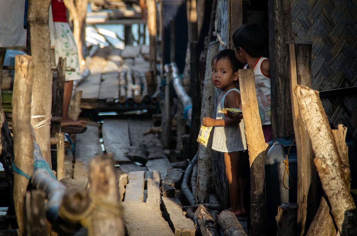 Two children stand near a wooden walkway in a rustic setting, with one child looking into the distance and holding a snack bag, as they dream of future island hopping adventures.