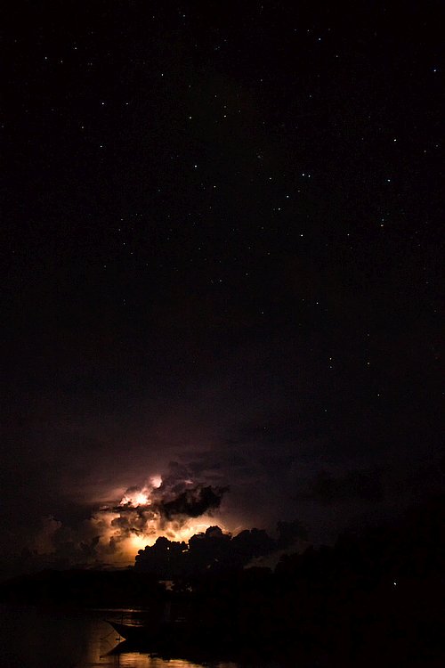 A dark night sky filled with stars, illuminated by distant lightning behind clouds, reflects softly on a calm body of water below, setting the perfect scene for an unforgettable boat tour.