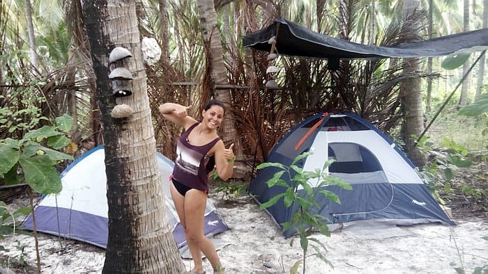 A smiling person in a swimsuit gives a thumbs-up in a forest campsite, capturing the essence of island hopping in Palawan. Two tents set up under a tarp among palm trees create the perfect base for adventure, with sandy ground and leaves completing the idyllic scene.