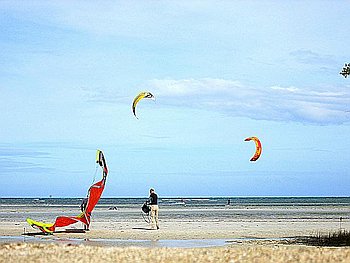 A beach scene with a person strolling on the sand near vibrant kiteboarding sails. The sky is clear with a few clouds, and the ocean stretches in the background, inviting adventurous souls to explore island hopping delights.