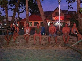 A group of six people sit on a low wall under trees during twilight, discussing their recent kitesurfing adventures. Behind them, buildings glow with lights, and summer attire hints at the day's beach activities. The sandy ground enhances the coastal vibe of this serene setting.