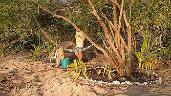 A person enjoys a serene camping spot in Palawan, standing on a sandy area surrounded by trees. Nearby, a tent and hammock blend into the tranquil setting with plants arranged in a circular pattern bordered by rocks. A blue bucket and chair complete this idyllic outdoor retreat.