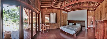 A cozy Palawan bedroom with wooden walls and ceiling, featuring a double bed, a wooden desk with chairs, and a window with curtains. Enjoy views of the outdoors through sliding glass doors to the left, perfect after a day of island adventure or boat tours.