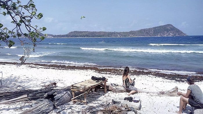 A scenic beach view features a sandy shore with scattered seaweed, two people relaxing near a rustic wooden bench, and ocean waves under a clear blue sky. An island, perfect for island hopping, is visible in the distance.