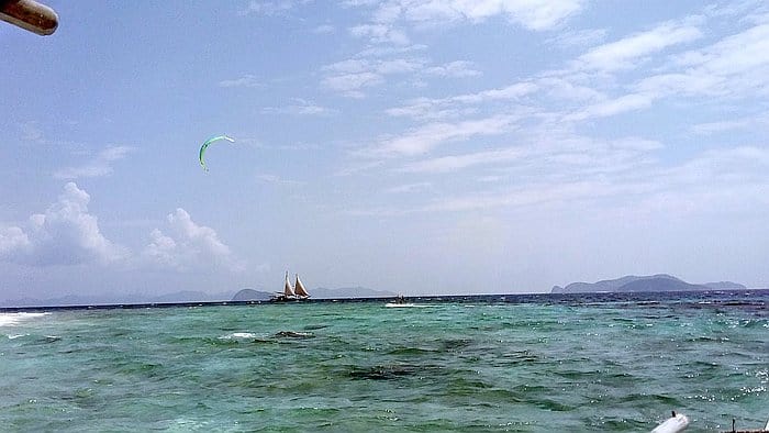 A scenic view of the ocean with clear blue-green water, a distant sailboat, and a kite surfer in the sky under a partly cloudy sky. Islands beckon on the horizon, perfect for island hopping adventures or leisurely boat tours.