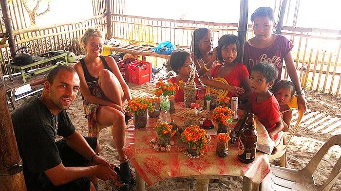 People sitting around a table with drinks and flowers in a bamboo hut, perhaps sharing tales from their recent boat tour. Several adults and children are smiling and looking at the camera, radiating joy and warmth.