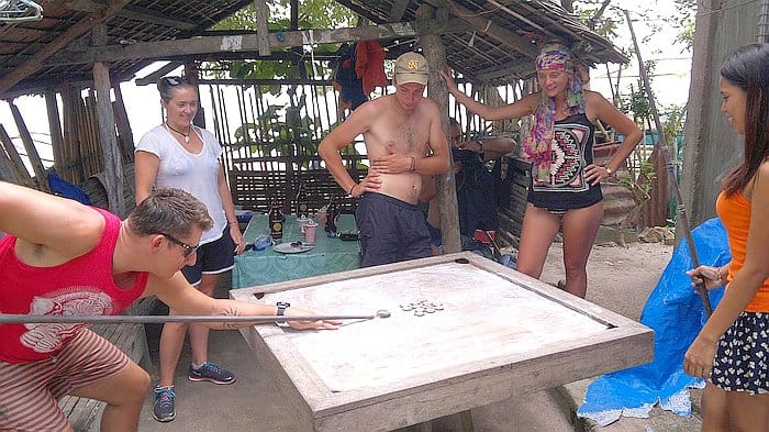 A group of people plays a game around a wooden table outdoors, under a rustic shelter, sharing laughs as tales from their earlier boat tour add an adventurous twist to the friendly competition.