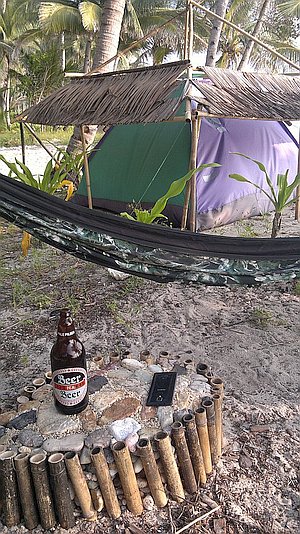 A tropical campsite with a hammock, tents, and a bamboo table holding a beer bottle and a phone, surrounded by palm trees. Nearby, whispers of an upcoming boat tour promise an adventure just beyond the shoreline.