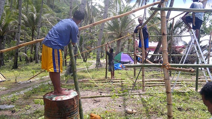 People building a bamboo structure outdoors, surrounded by palm trees and grassy areas, resemble explorers prepping for a distant boat tour, with a tent pitched in the background.