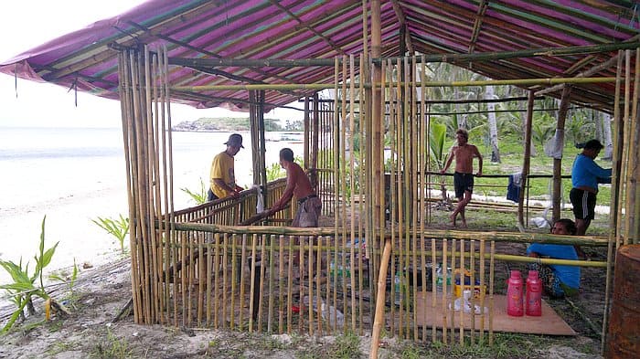 People constructing a bamboo structure on the beach, with the sea visible in the background, pause to wave at a nearby boat tour passing by.