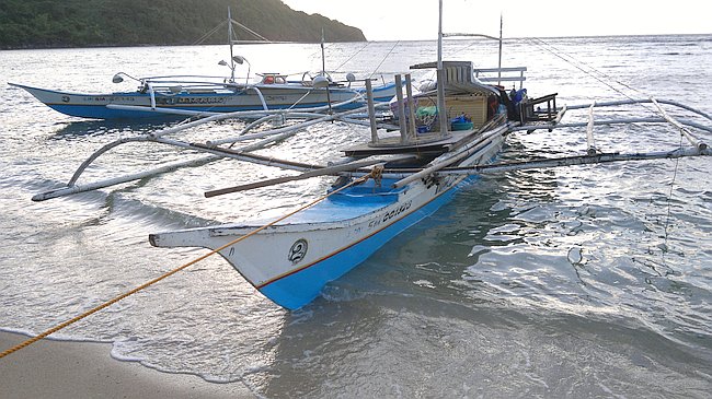 Two traditional outrigger boats, perfect for a serene boat tour, are anchored at the shoreline. The scenic hilly backdrop and gentle waves lapping at the beach complete this idyllic setting.