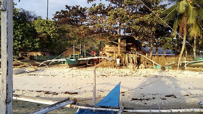 Boats rest on a sandy beach, with trees and a rustic wooden house providing the perfect backdrop under a clear sky—an idyllic starting point for any enchanting boat tour.