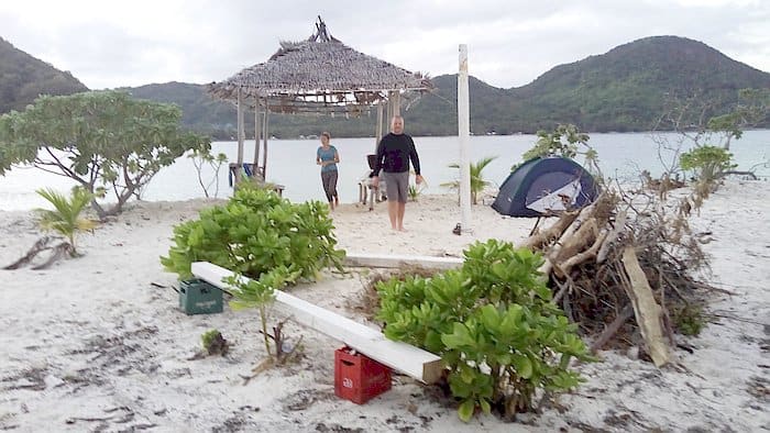 A beach campsite with a thatched shelter, tent, and two people. Greenery surrounds the area, with hills and water in the background. Nearby, a boat tour offers a chance to explore the serene waters beyond the shore.