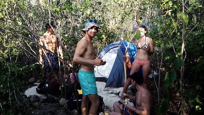 Four people in swimsuits set up camp near a blue tent in a forested area, surrounded by trees and dappled sunlight, perfecting their island hopping itinerary after an exhilarating boat tour.