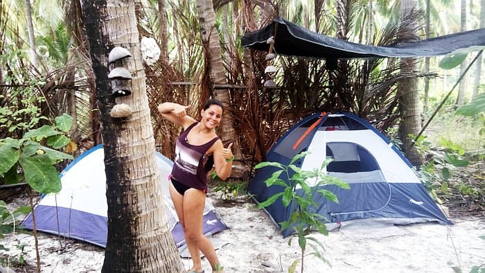 A person in a swimsuit is smiling and posing next to tents in a tropical campsite with palm trees, ready for an adventurous day of island hopping.