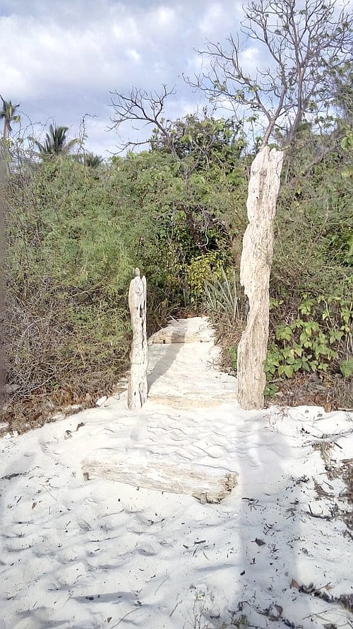 A sandy path leads into lush greenery with two tall, weathered wooden posts on either side under a cloudy sky, inviting travelers to embark on an island hopping adventure.
