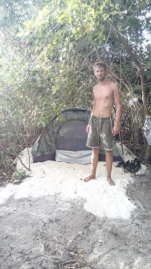 A shirtless man stands by a camouflaged tent on a sandy ground surrounded by trees, taking a break from his island hopping adventure.