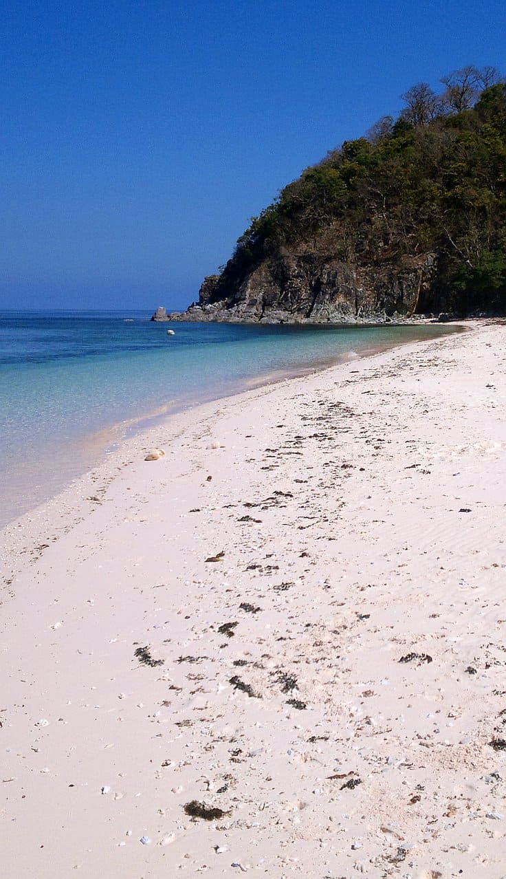 A pristine beach with white sand and clear blue waters stretches alongside a lush, green hillside. The sky is clear and bright, creating a serene tropical atmosphere perfect for island hopping or leisurely boat tours.