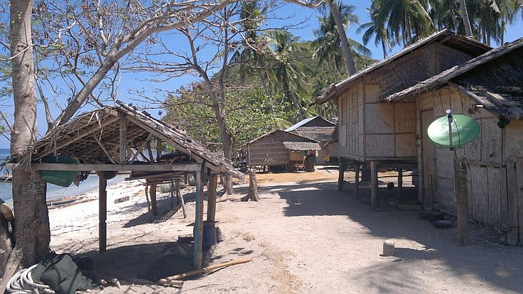 A serene beach scene with several wooden huts nestled among palm trees awaits. The sandy path lined with dry branches leads to the blue sea, perfect for island hopping in the background under a clear sky. A green satellite dish is attached to one hut.
