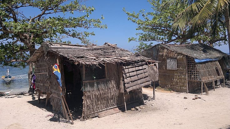 Two rustic thatched huts on a sandy beach, shaded by palm trees. A traditional boat awaits island hopping adventures on the shoreline, with the blue ocean in the background. Bright, sunny day with clear skies.