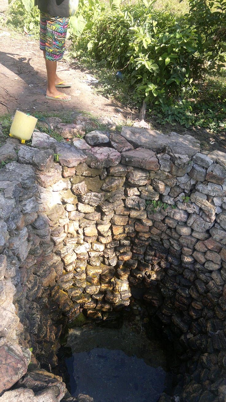 A person stands near an open, stone-lined well with clear water at the bottom. Greenery surrounds the area, and a yellow container is placed on the edge of the well. Nearby, whispers of island hopping adventures and boat tours beckon from beyond leafy horizons.