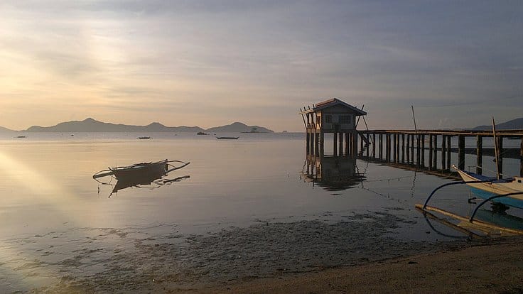 A serene sunset over calm waters features a wooden pier extending into the sea, a small stilt house at the end, and a traditional boat resting on gentle waves, perfect for island hopping. Distant islands beckon adventurers to explore on enchanting boat tours.