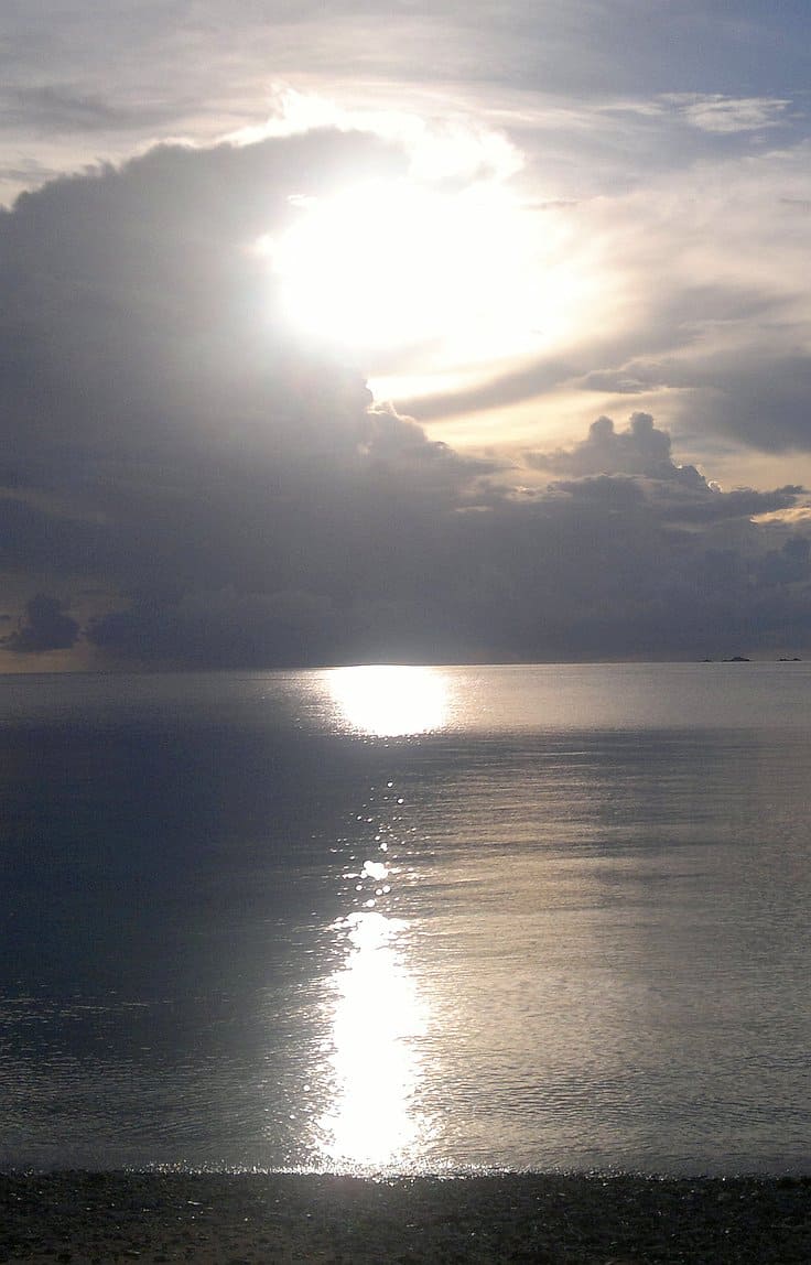 Sun shining through clouds over a calm sea, creating a shimmering reflection on the water and casting a serene atmosphere, perfect for boat tours. The horizon is visible with soft light and gentle waves, inviting the thought of island hopping adventures ahead.