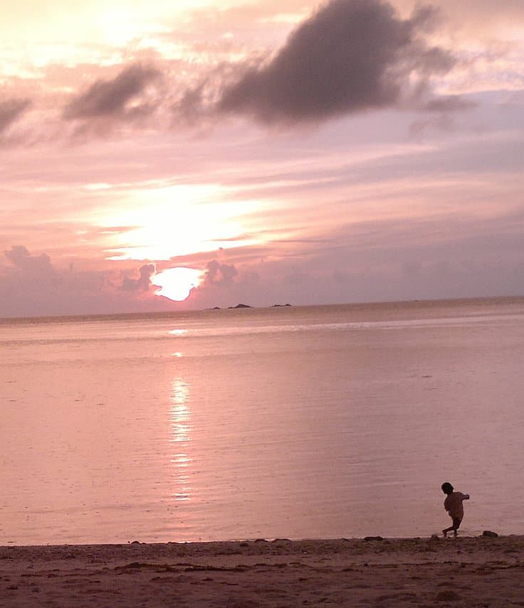 A serene beach scene at sunset with a young child playing near the water's edge, dreaming of future island hopping adventures. The sky and sea are tinted with soft pink and orange hues, while scattered clouds add texture to the tranquil scene.