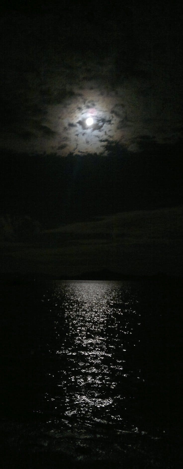 A moonlit night sky with clouds partially obscuring the moon enhances the mystique of island hopping. Its light reflects across a dark, rippled body of water, creating a shimmering trail on the surface as if guiding boat tours through an enchanting nocturnal journey.