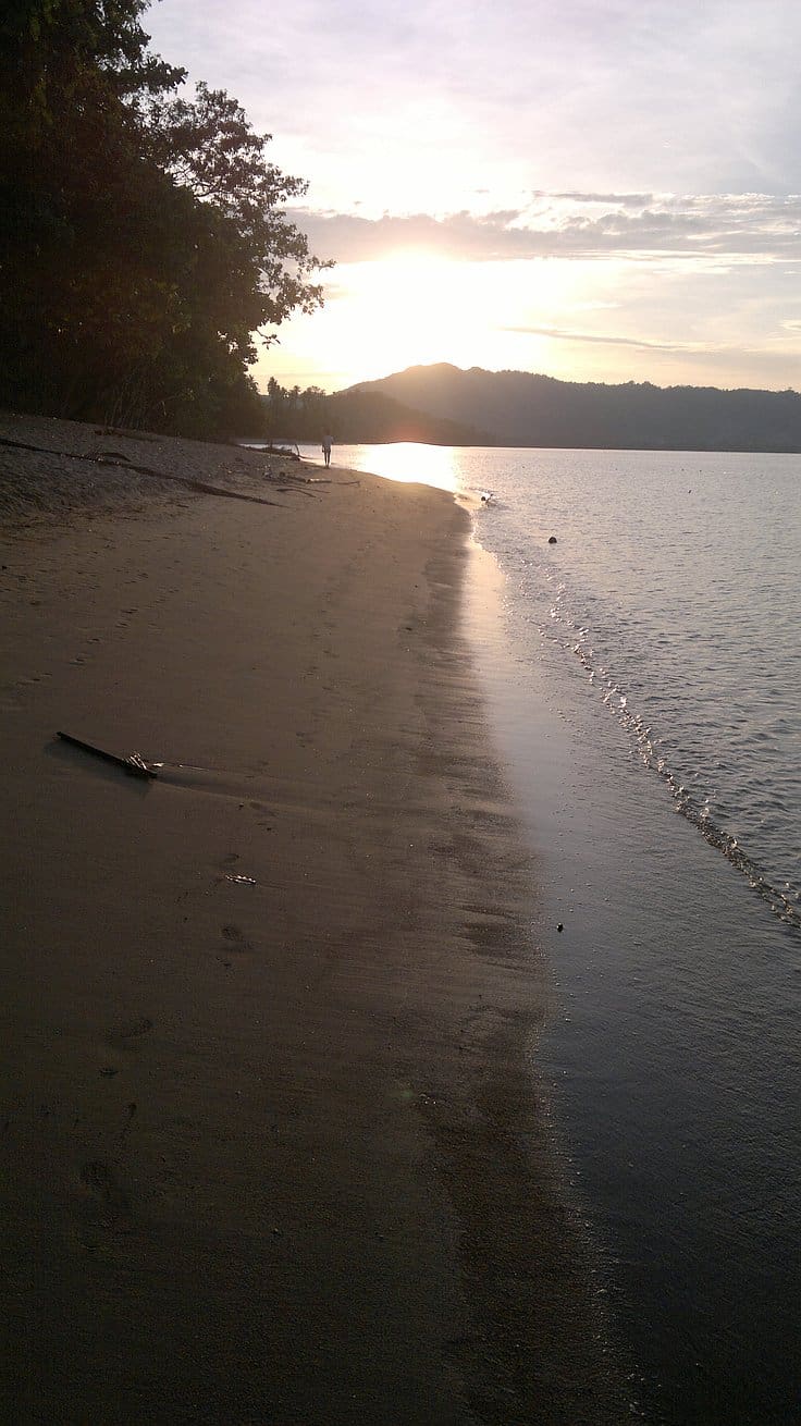 A tranquil beach scene at sunrise, the sun partially obscured by distant hills. Gentle waves touch the sandy shoreline as silhouettes of trees stand on the left, hinting at nearby island hopping adventures and serene boat tours awaiting discovery.