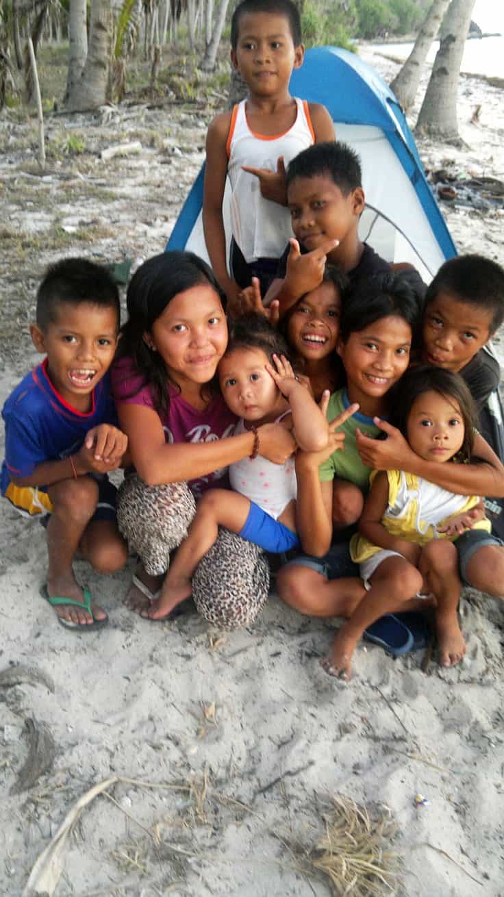 A group of children posing and smiling in front of a blue and white tent on a sandy beach with palm trees swaying gently in the background, capturing the joyful spirit of island hopping. They are sitting and kneeling, looking cheerful and relaxed.
