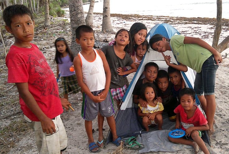 A group of children gathered on a sandy beach near a small blue and white tent, taking a break from their island hopping adventure. Some are standing, while others are seated inside the tent. Palm trees and the ocean are visible in the background, with everyone looking happy and relaxed.