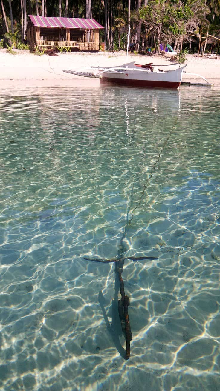 A small wooden boat is anchored in clear, shallow turquoise water near a sandy beach, perfect for island hopping. A rustic wooden hut stands beneath palm trees in the background, creating a tranquil, tropical scene.