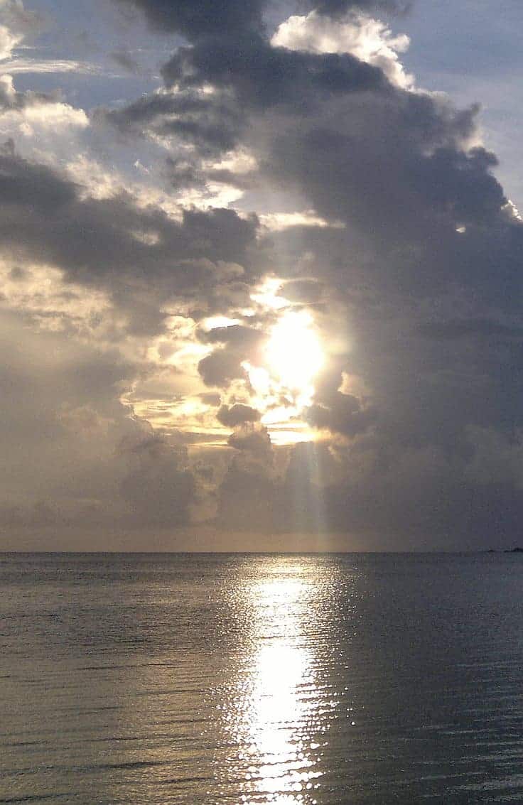 A serene seaside scene with the sun setting behind thick clouds, casting a radiant glow and shimmering reflection across calm ocean waters. The sky is a blend of gray and golden hues, perfect for island hopping adventures or tranquil boat tours.