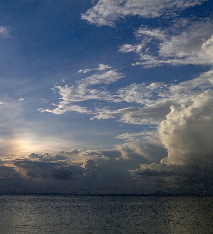 A serene seascape at sunset, with the horizon meeting the calm ocean. The sky is partly cloudy, featuring streaks of light and patches of blue, as a boat tour gently glides past, enhancing the tranquil atmosphere while the sun sets behind distant clouds.