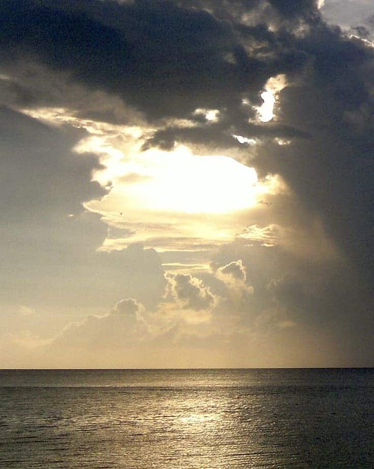 A dramatic seascape at sunset, with sunlight piercing through dark clouds, reflecting off the calm water as boats meander by. The sky’s hues of orange, gray, and gold create a serene atmosphere, perfect for island hopping adventures.