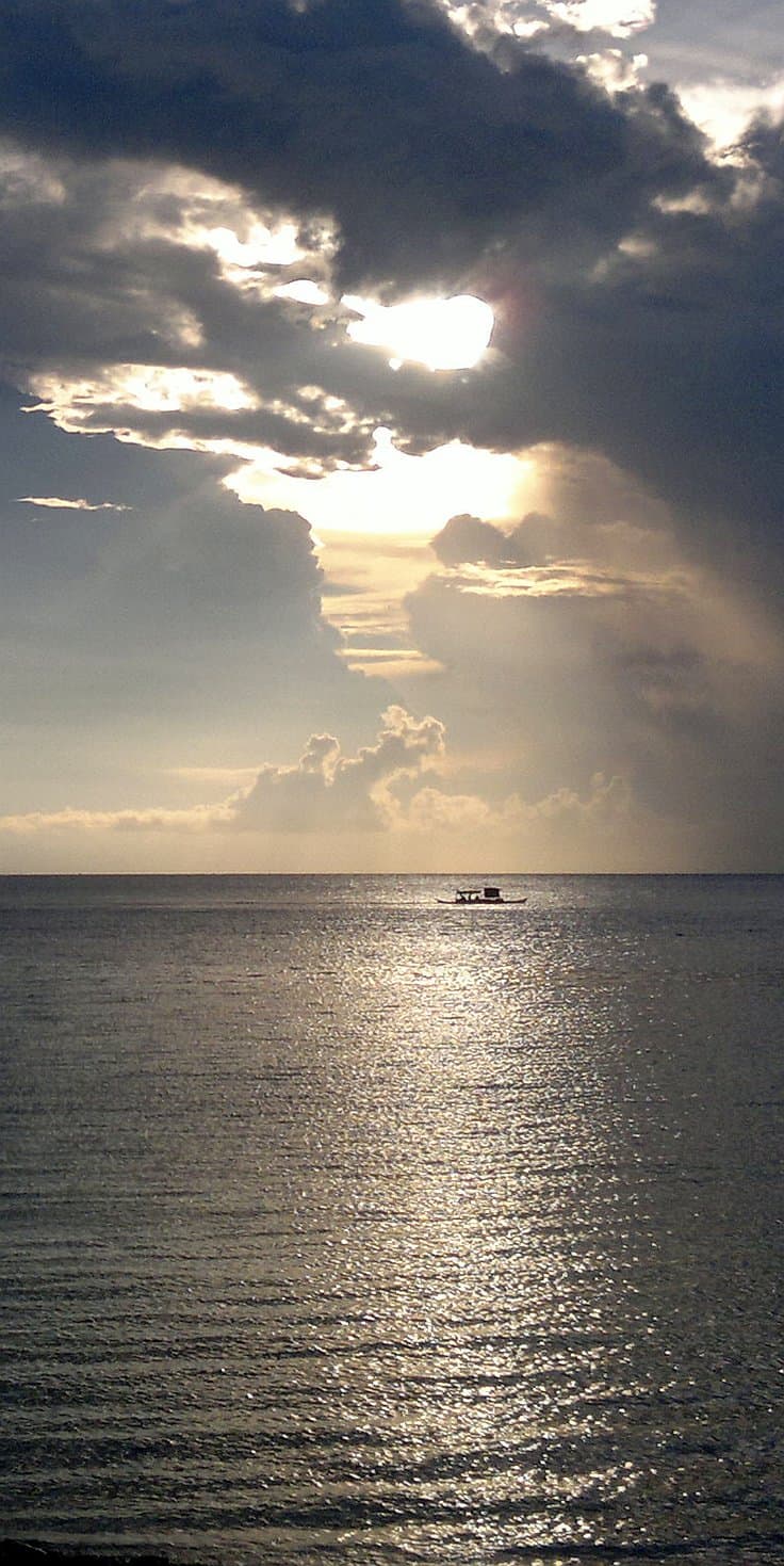 A small boat from a local island hopping tour floats on a calm sea under a dramatic sky with the sun partially obscured by clouds, casting a shimmering reflection on the water.