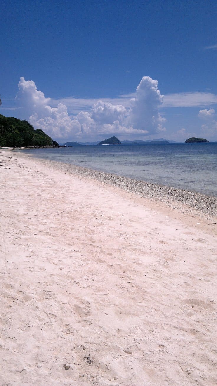 A tranquil beach scene with a clear blue sky, fluffy clouds, and calm ocean waves offers the perfect setting for island hopping. White sand stretches along the shore, with green hills visible in the background, inviting visitors to explore nearby islands through serene boat tours.
