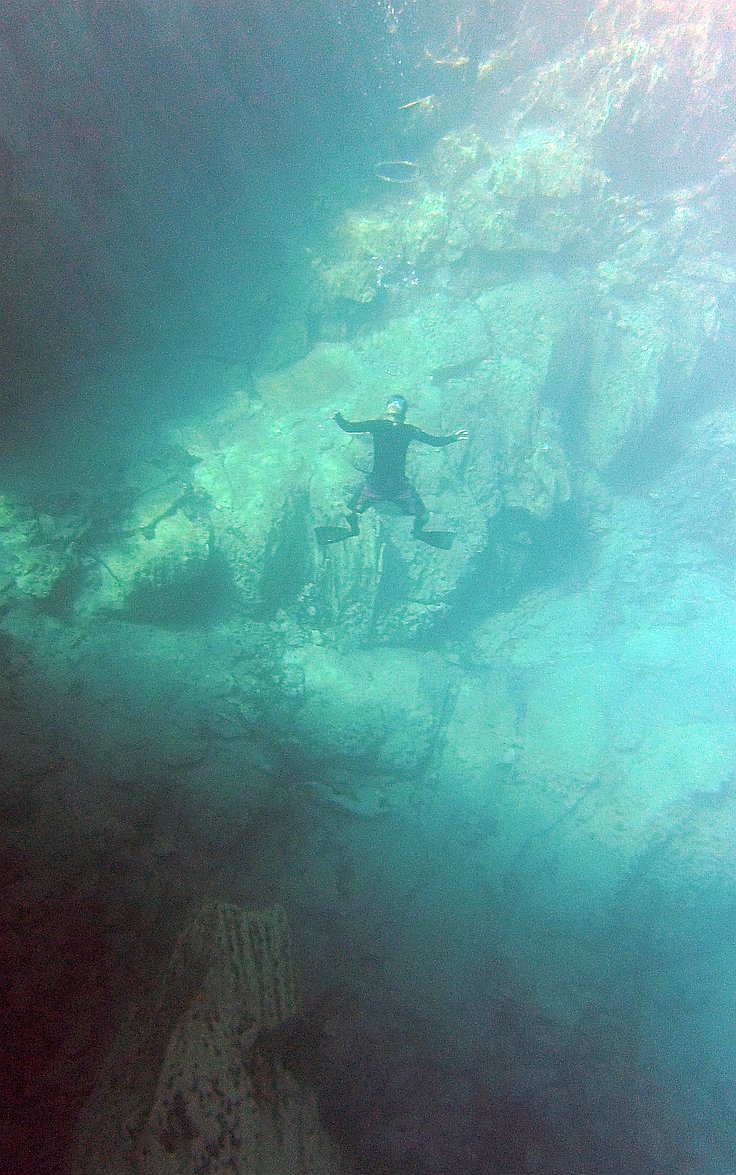 A person in a wetsuit floats underwater with arms and legs spread out, surrounded by clear blue water and rock formations. Sunlight filters through the surface, hinting at nearby island hopping adventure tours just above.